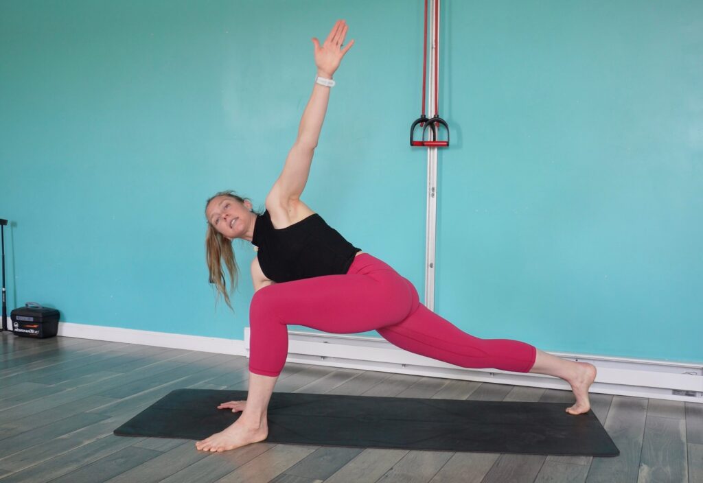 Dr. Chloe with her left leg forward, right leg back, pushing through her toes in a deep lunge. Her right hand is on the ground and shes twisting open from her trunk with her left fingertips pointing towards the ceiling as a trail running stretch